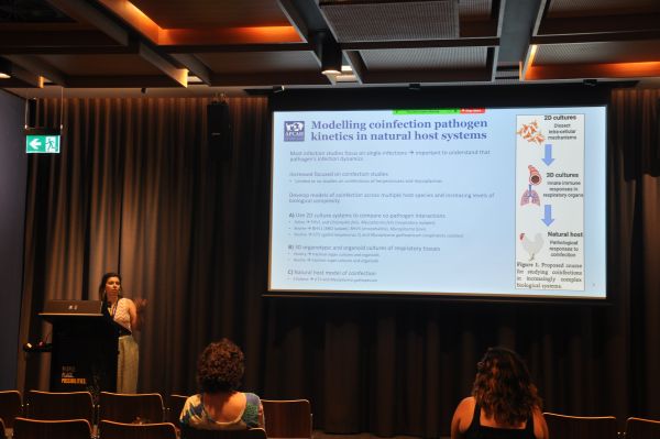 A speaker at the podium next to a slide title 'Modelling coinfection pathogen kinetics in natural host systems'