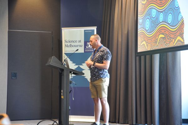 A speaker at the podium; a corner of their slide and a 'Science at Melbourne' banner are visible