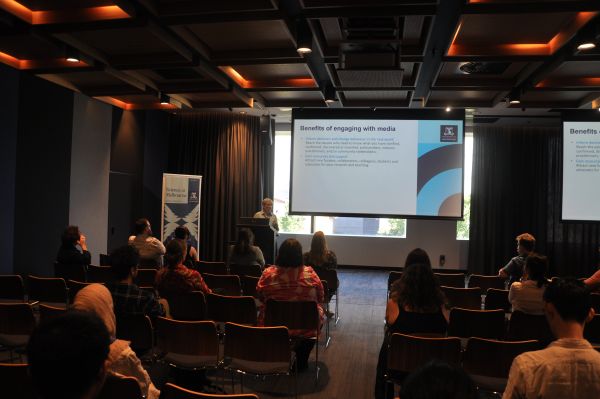 A speaker at the podium next to a slide titled 'Benefits of engaging with media'. Most of the audience is also in frame