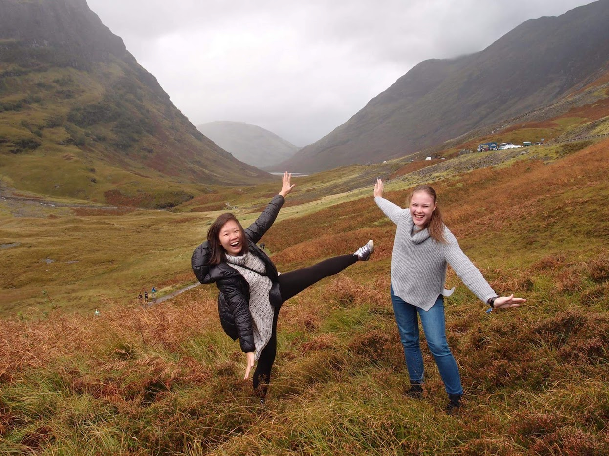 Renee and a friend posing playfully in a large valley. Mist-shrouded mountains rise in the background.