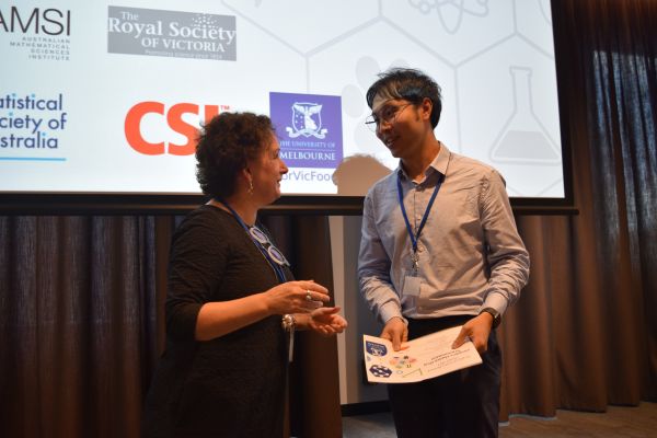 Moira O'Bryan presenting a certificate to a speaker; the bottom of a slide is visible in the background