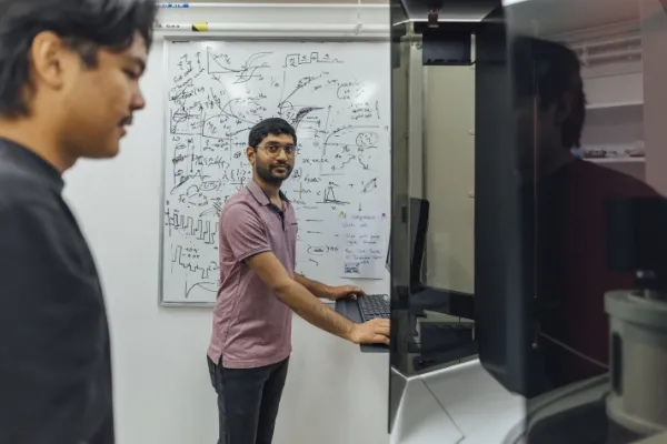 One man standing in front of a whiteboard covered in equations, looking back at another person standing at a monitor.