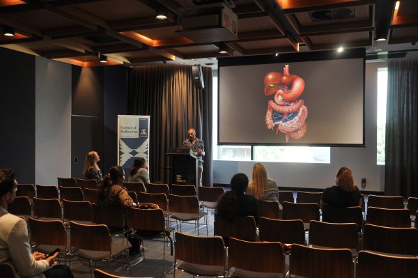 A speaker at the podium next to a slide showing a cartoon of the human digestive system. Several rows of the audience are also visible