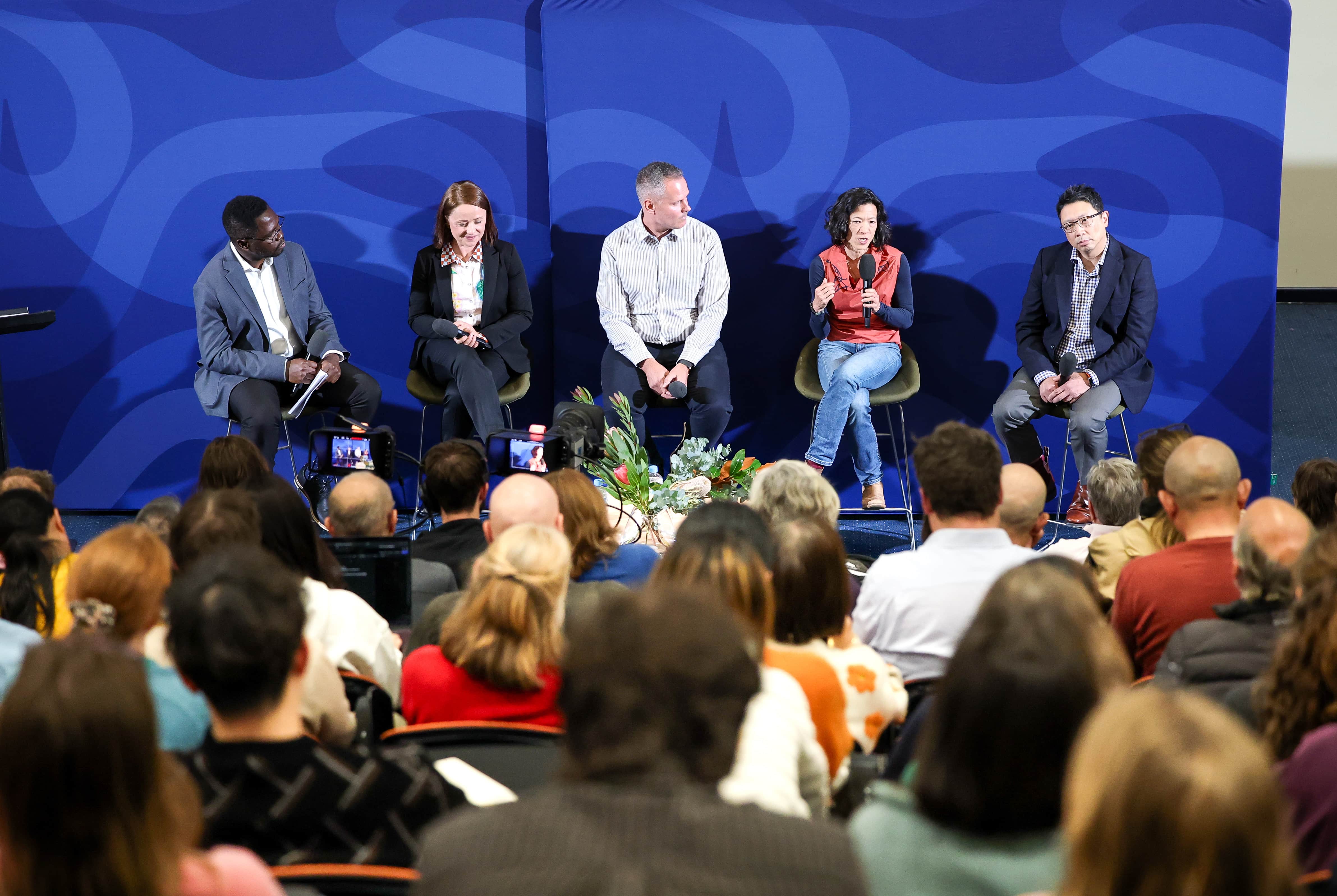 Five panel members sit on a stage facing an audience