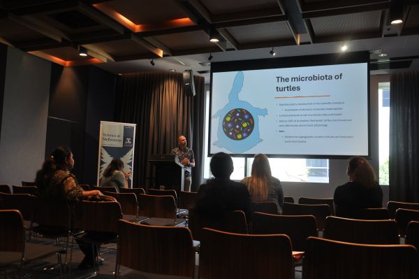 A speaker at the podium next to a slide titled 'The microbiota of turtles'. Sseveral rows of the audience are also visible