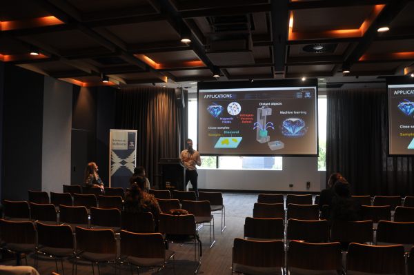 A speaker at the podium next to a slide titled 'Applications'; several rows of the audience are visible