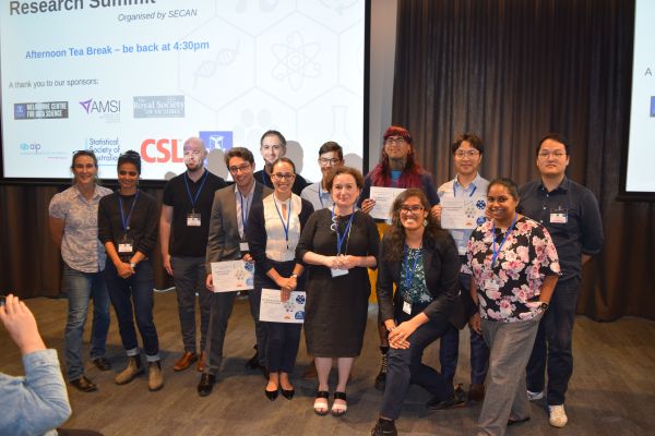 Speakers with certificates, conference organisers and Moira O'Bryan posing for the camera; most of a slide can be seen in the background