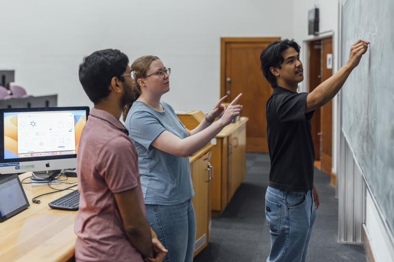 Student writes on blackboard while two other students watch and give instructions
