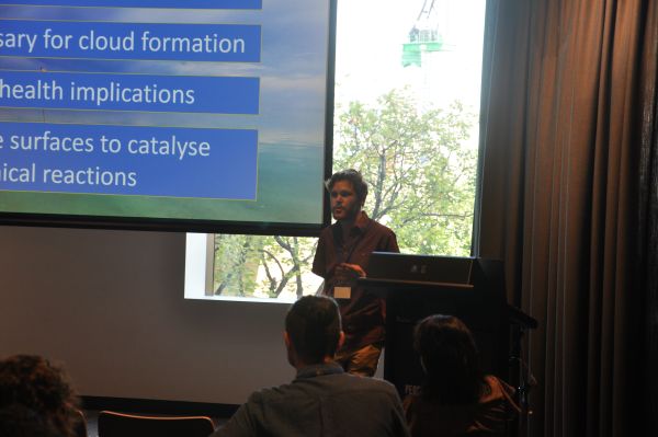 A speaker at the podium; one corner of the slide is visible
