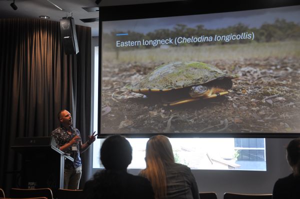 A speaker at the podium next to a slide titled 'Eastern longneck (Chelodina longicollis)', containing a photo of a turtle