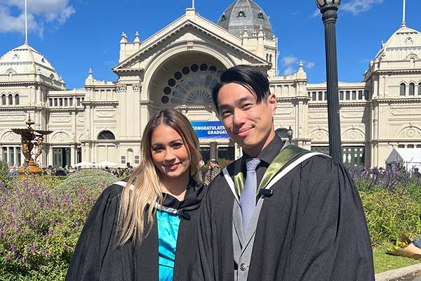 Two students in graduation gowns standing in front of the Exhibition Building