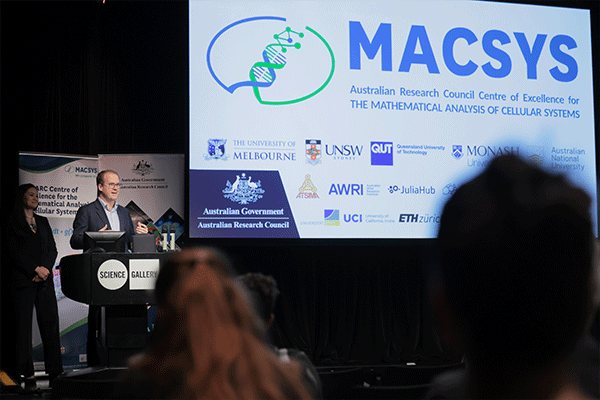 Professor Mark Cassidy standing at a lectern; behind him is a large screen displaying the MACSYS logo