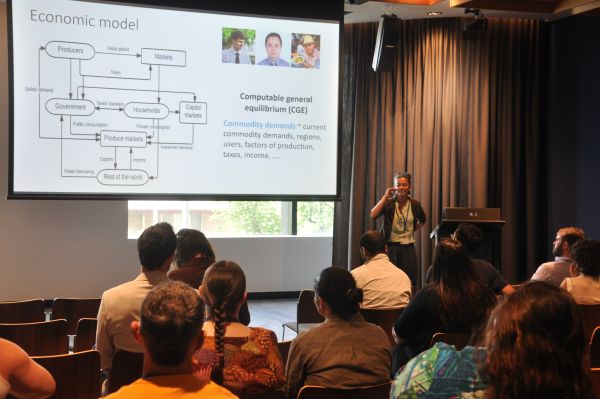 A speaker at the podium next to a slide showing a flowchart with the title 'Economic model'