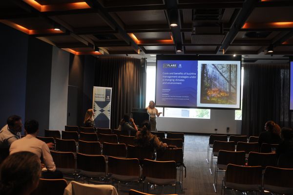 A speaker at the podium next to a slide titled 'Costs and benefits of bushfire management strategies under a changing climate and environment'. Several rows of the audience are in frame