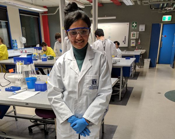 A picture of Vandana in a white lab coat, wearing blue gloves and goggles, in a lab with other students for one of her Biotechnology classes