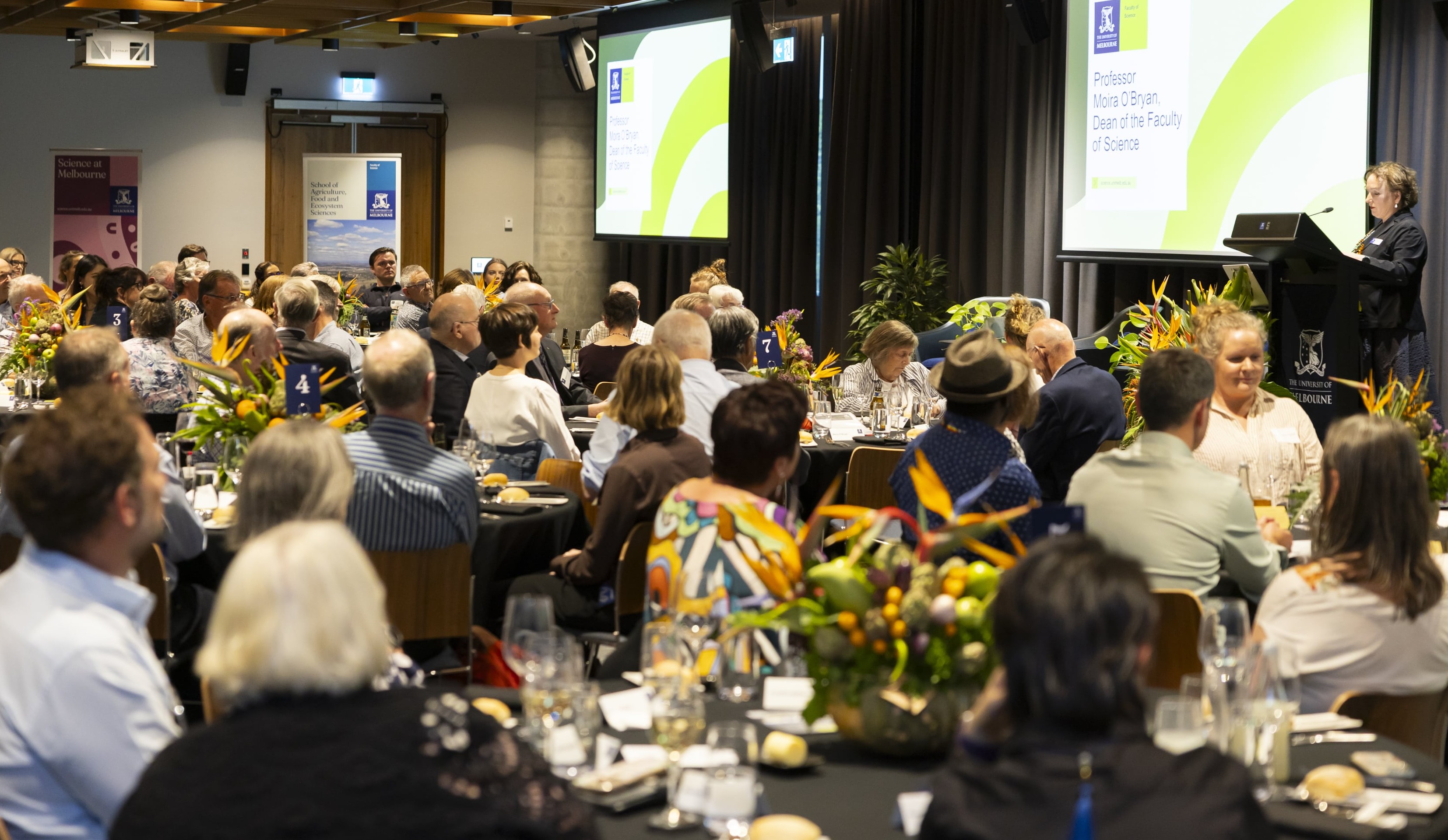 A crowd of people seated at tables in a large function room are addressed by a woman at a lectern