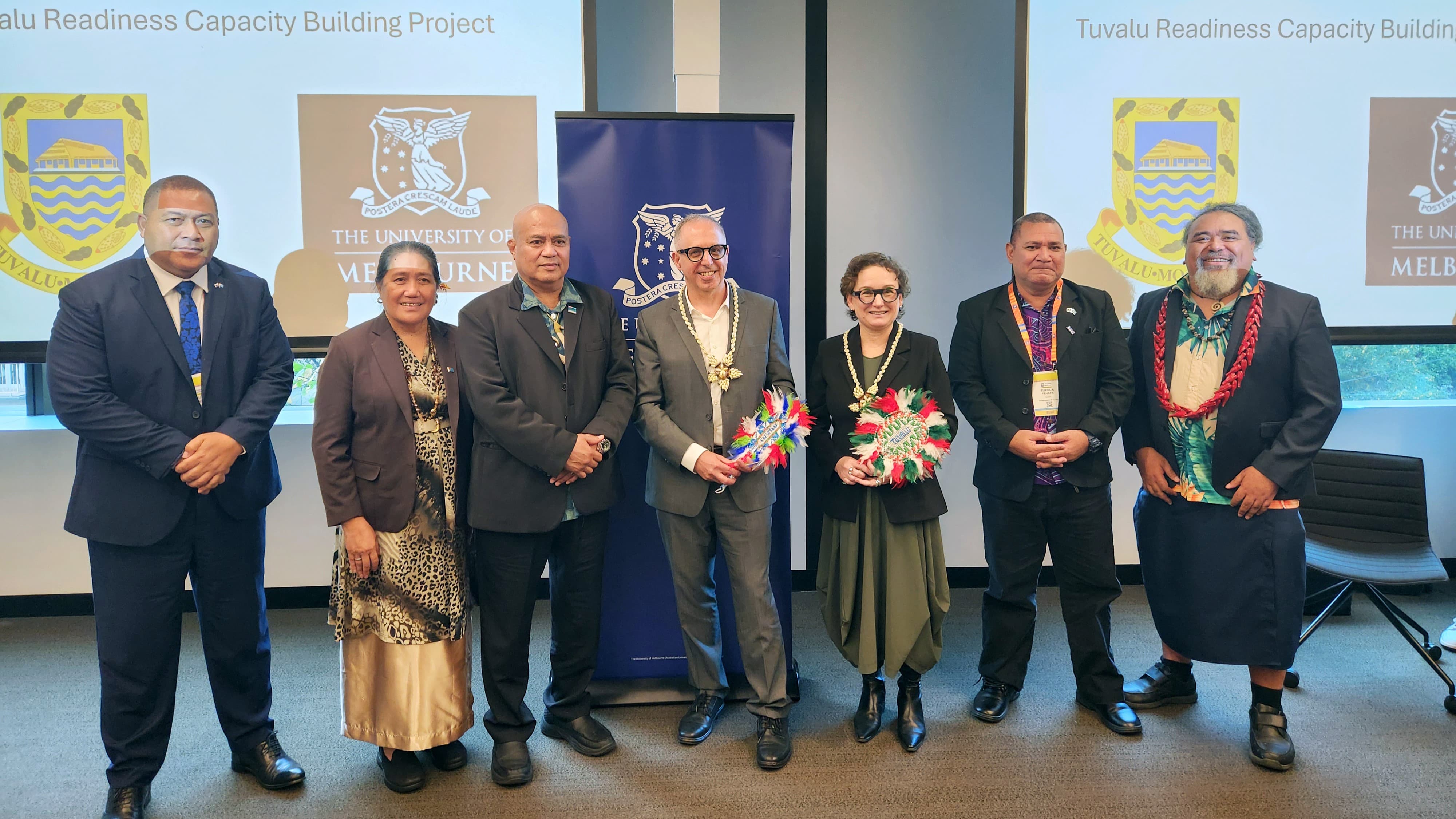 Seven Australian and Tuvaluan people stand side-by-side in a room. A projected slide in the background reads "Tuvalu readiness capacity building project"