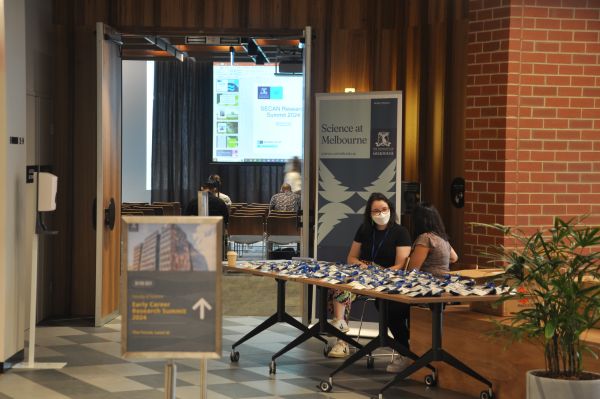 Two summit organisers speaking at the registration desk; slides and some of the audience in the presentation room can be seen to the side