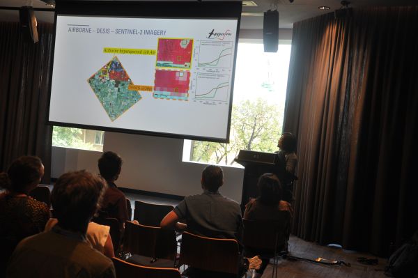 A speaker at the podium next to a slide titled 'AIRBORNE - DESIS - SENTINEL-2 IMAGERY'. Several rows of the audience are also visible