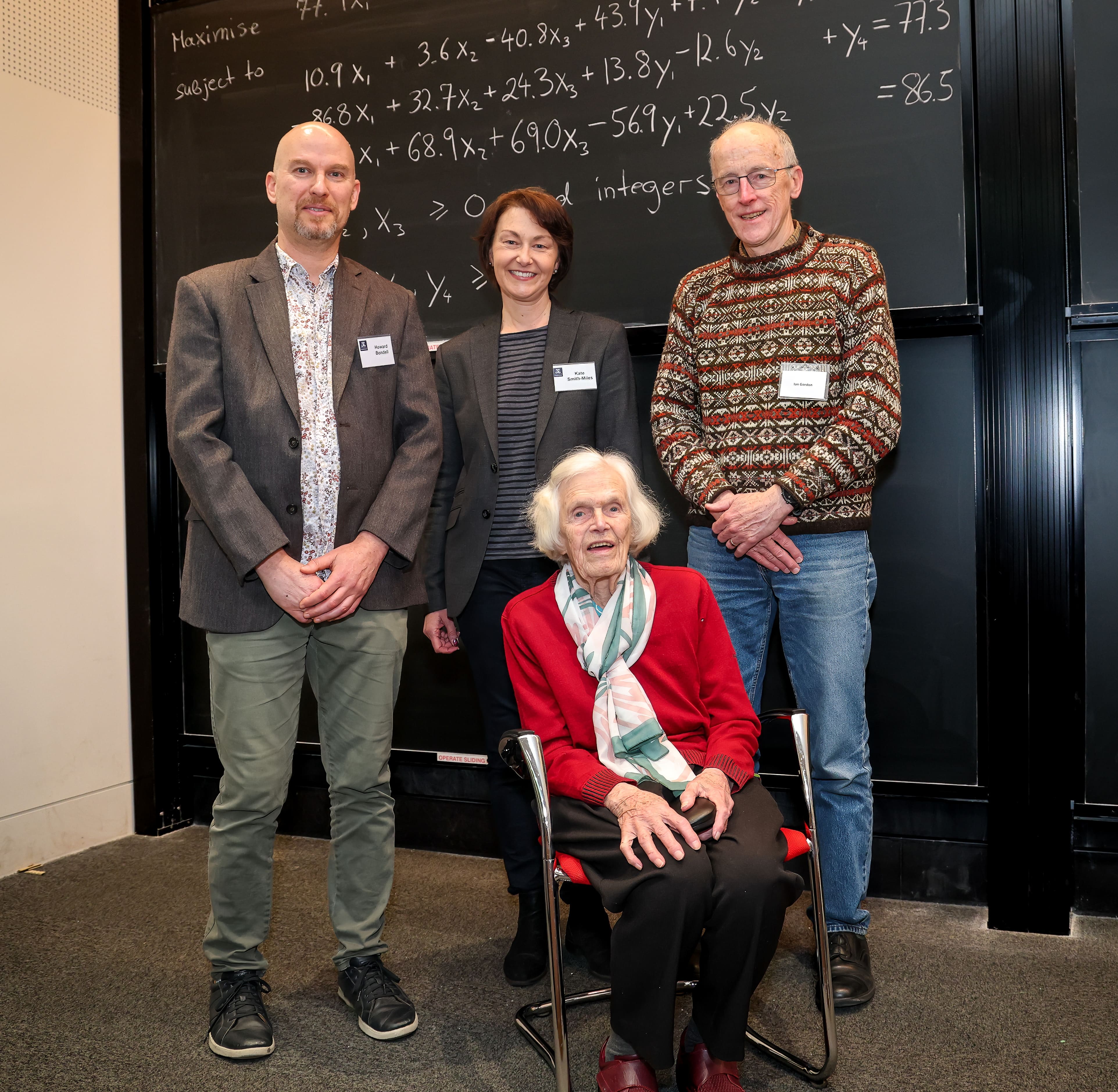 Three people stand and one sits in front of a blackboard covered with mathematical symbols