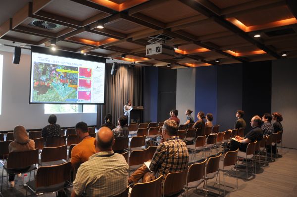 A speaker at the podium next to a slide titled 'AIRBORNE - DESIS - SENTINEL-2 IMAGERY'. Most of the audience is visible