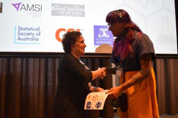Moira O'Bryan presenting a certificate to a speaker; the bottom of a slide is visible in the background