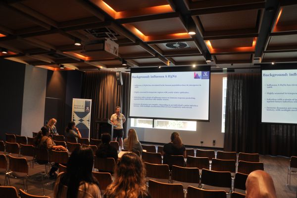 A speaker at the podium, next to a slide titled 'Background: Influenza A H3N2'. Several audience members are also visible
