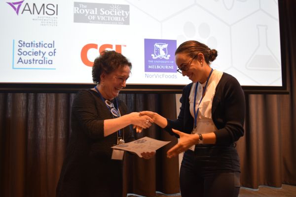 Moira O'Bryan presenting a certificate to a speaker; the bottom of a slide is visible in the background