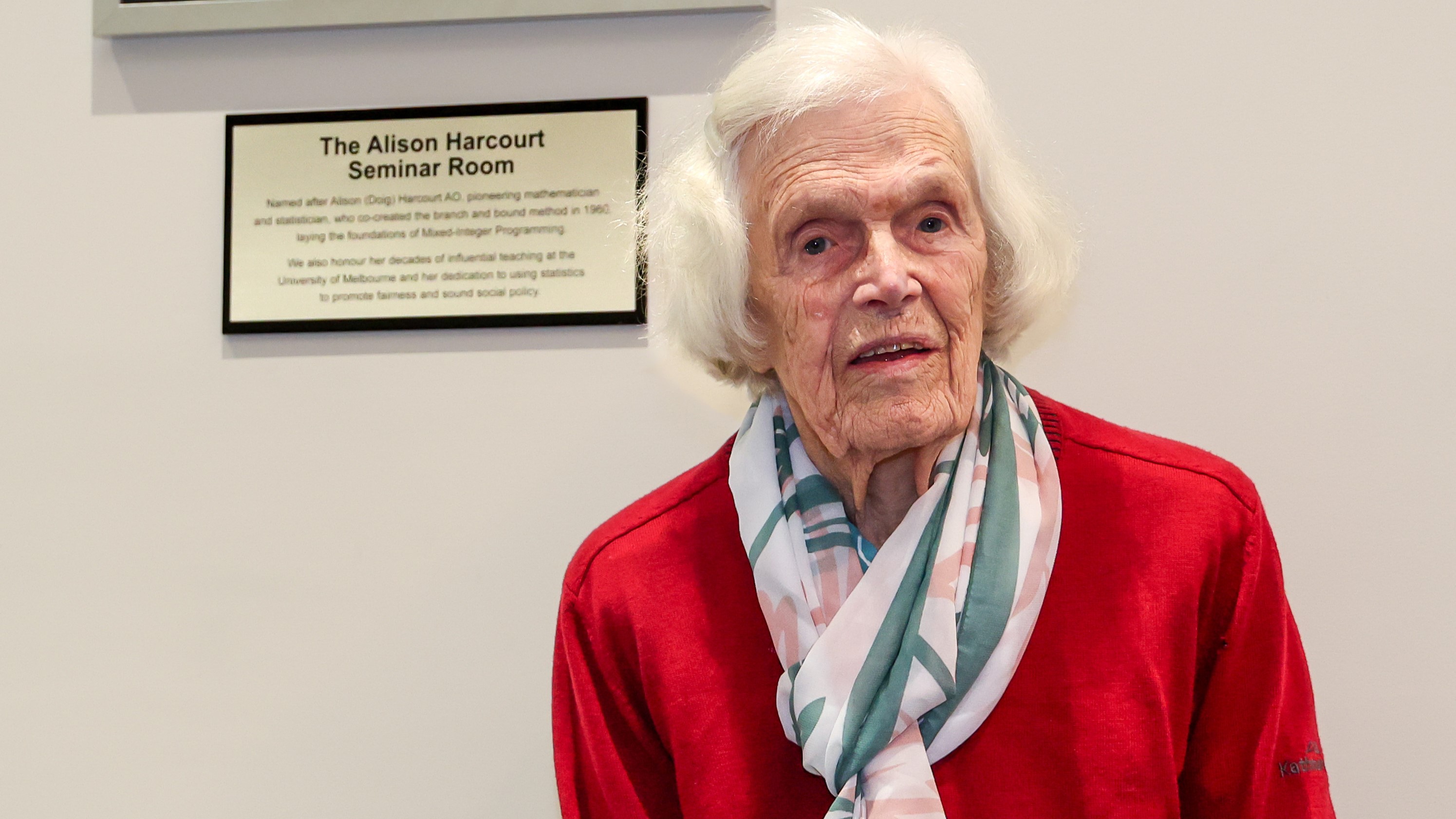A woman (Alison Harcourt) stands in front of a wall plaque stating that the room is named in her honour